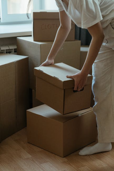 A person wearing a white shirt and light-colored pants is in an indoor space, handling a cardboard box during a house relocation process. The individual is grasping the box firmly, positioning it on top of a stack of other unopened cardboard boxes, all sealed with packing tape. Several additional boxes of varying sizes, some labelled with words such as 'Clothes,' are visible nearby, indicating packing preparations. The scene shows a well-lit room with natural light coming through a window, and the person appears to be in the midst of a packing or moving activity, which is part of the furniture transport and home relocation services offered by Man and a Van Hammersmith. The focus on packaging materials and careful handling reflects the packing and moving logistics involved in professional removals, emphasizing precise and secure packing for a smooth transport process.