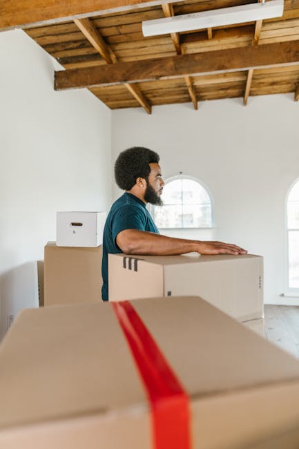 A man with dark curly hair and a beard, wearing a dark blue T-shirt, is seen in a spacious property with white walls and a wooden ceiling with exposed beams. He is standing behind several stacked card