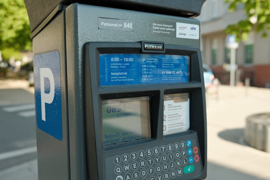 Close-up view of a parking meter situated on a city pavement with trees and residential buildings in the background. The meter displays parking rates and hours of operation on a blue-lit screen, with