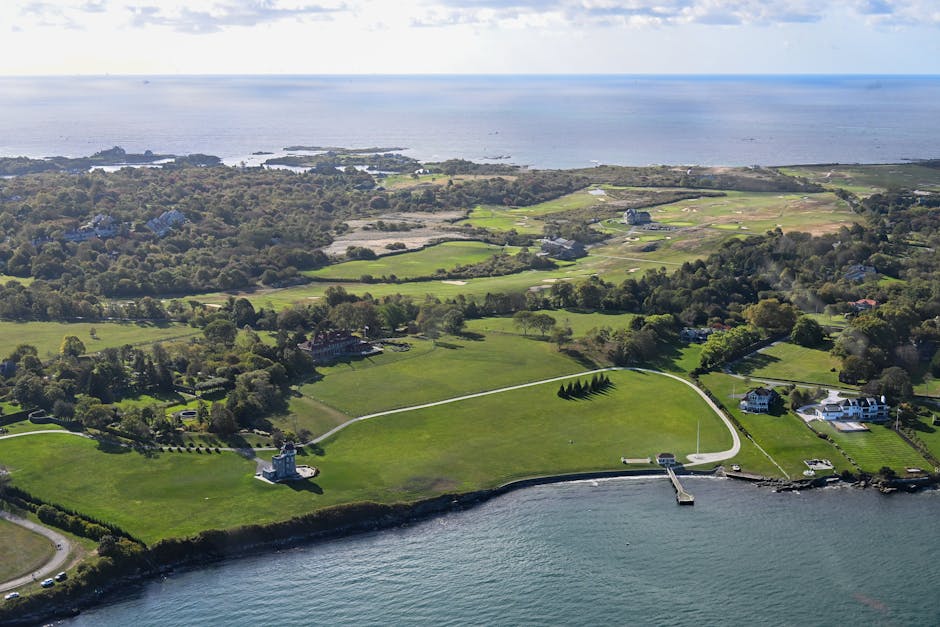 Aerial view of a coastal area featuring lush green lawns, several residential buildings, and a winding driveway leading to a lighthouse situated near the water's edge. The property is surrounded by mature trees and open fields, extending towards the shoreline and the sea in the background. The scene is illuminated by natural daylight, with clear visibility of the landscape's details. This image may relate to house or property relocations, highlighting the spacious outdoor environment and the nearby waterfront. The photograph captures the landscape in a professional manner, suitable for illustrating moving or relocation services such as those offered by Man and a Van Hammersmith, focusing on the characteristics of a scenic and accessible location for home relocations or furniture transport.