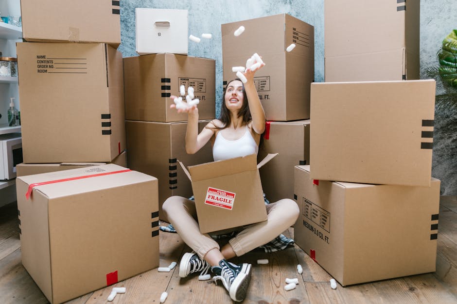 A woman sitting on the floor inside a room surrounded by numerous cardboard boxes, some labelled as fragile, while packing or preparing for a house relocation. She is wearing casual clothes with striped sneakers and has a happy expression as she tosses white packing materials into the air. The boxes vary in size, with some stacked on each other and others placed on the wooden floor, indicating an organized packing process. Behind her, a grey textured wall and a potted plant are visible, along with a doorway leading to another room. To the left, part of a white kitchen counter and a dispenser are seen. The environment is well-lit, and the scene reflects the packing and moving activities involved in home furniture transport and house removals, with [COMPANY_NAME] providing professional relocation services.