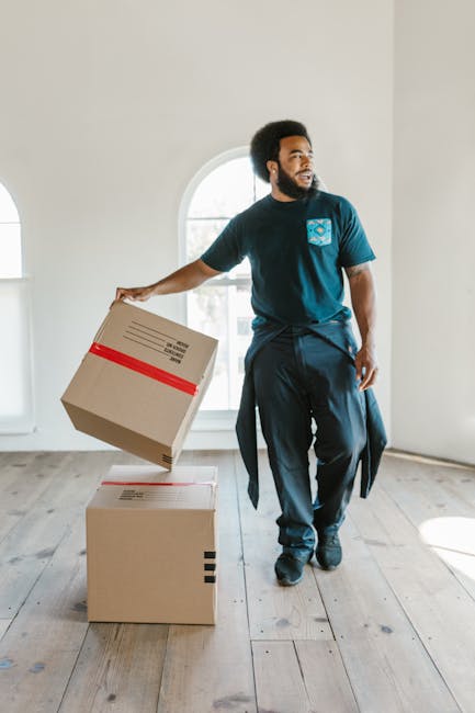 A young man with light skin, wearing a dark blue work jumpsuit and a black bandana with white patterns, is standing inside a room with a textured light grey wall. He is holding a clipboard and a blue pen, appearing to be noting details related to house removals. Surrounding him are cardboard boxes, one with a red and white label, and various packing materials like tissue paper or bubble wrap. To his left, there are two light blue mugs on a white shelf, alongside decorative items including a gold-colored object and a clear glass bottle. A large white potted plant with long green leaves is positioned on the top shelf above the mugs. The environment suggests a preparatory phase of a furniture transport or home relocation process, with the individual likely performing inventory or packing tasks as part of a professional removals service offered by Man and a Van Hammersmith.